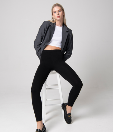 Woman wears Black Leggings, White Top, Grey Blazer and Black Brogue shoes. She sits on a white stool in a studio room.