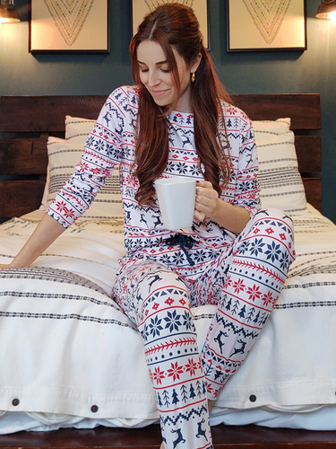 Woman wears the White Christmas Pyjamas whilst sitting on her bed with a hot drink. 