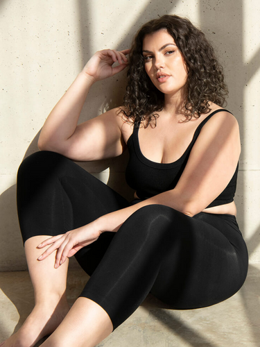 Woman wears black vest top and Cropped Black Everyday Leggings in Curve. She sits by a window, on the floor, with the shadows from the window frame reflecting onto her.