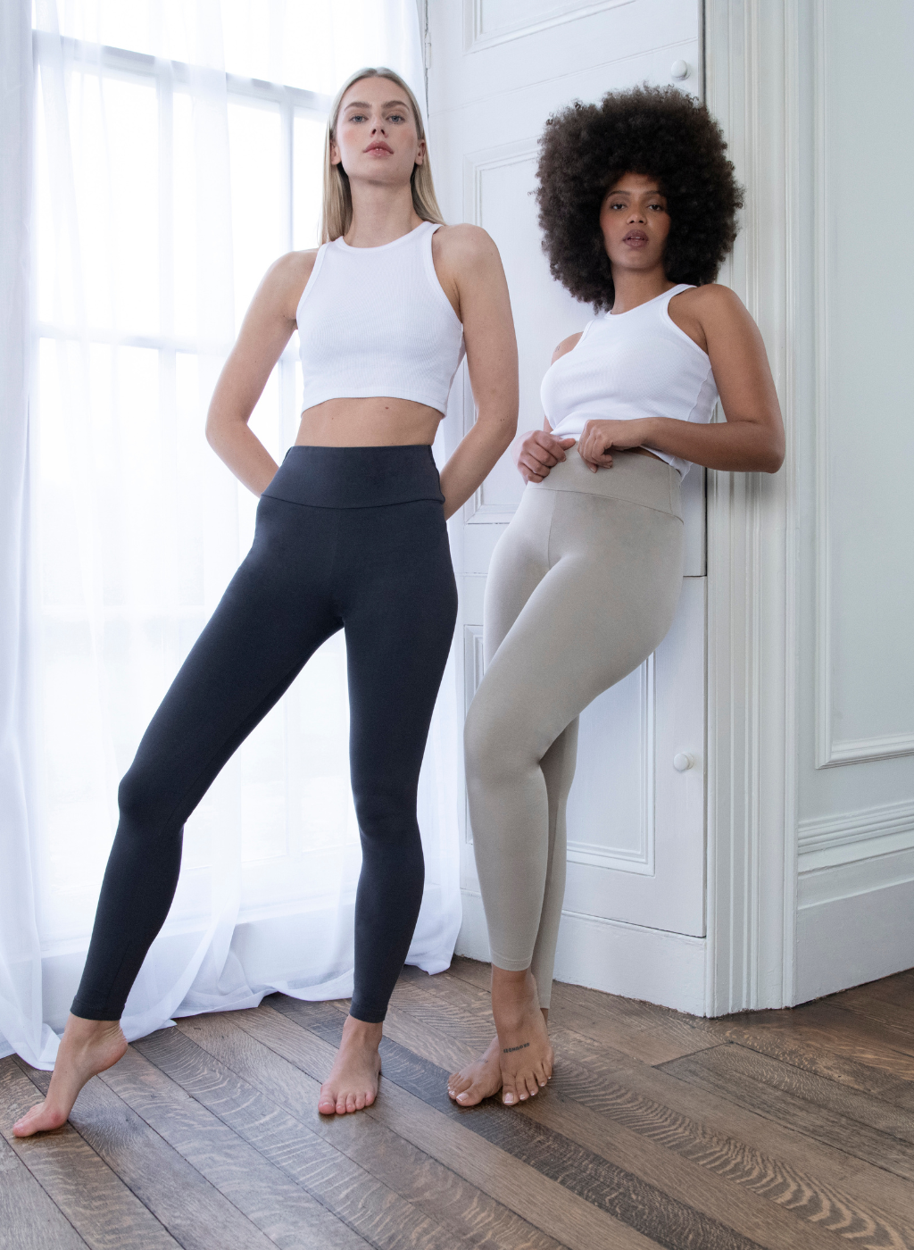 Two Model wearing the Everyday High Waisted Legging In colours Infinity Blue Marl and Oatmeal Beige. They are standing indoors, next to a white window and one model is leaning against a white wall. 