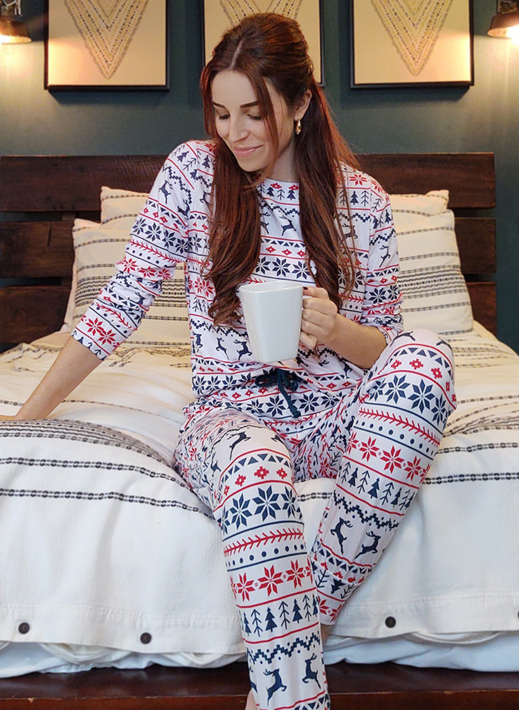 Woman wears the White Christmas Pyjamas whilst sitting on her bed with a hot drink.